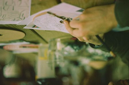 A hand scratches grooves into a clay disk. They're at an art table with supplies scattered on it.