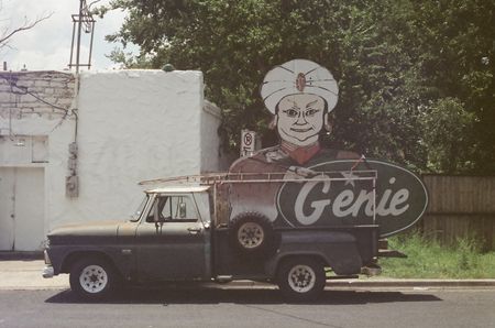 A vintage pickup truck is parked on the side of a street in Austin, TX. In the bed is a large sign with a genie illustration and the word Genie in a vintage script.