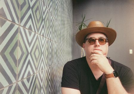 Joel Goodman, wearing a straw hat and tinted glasses, standing thoughtfully beside a geometric-tiled wall