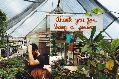Two people browsing plants in a plant shop with a sign overhead that reads Thank you for being a person.