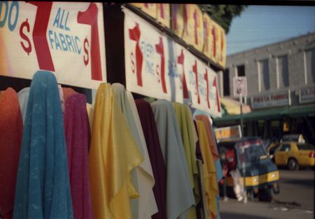 A row of colorful fabric with the same sign repeated above them. It reads: All Fabric $1. A LA street in the background. A yellow taxicab is parked there.